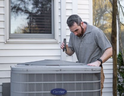 An HVAC technician inspecting an outdoor condenser unit with a flashlight