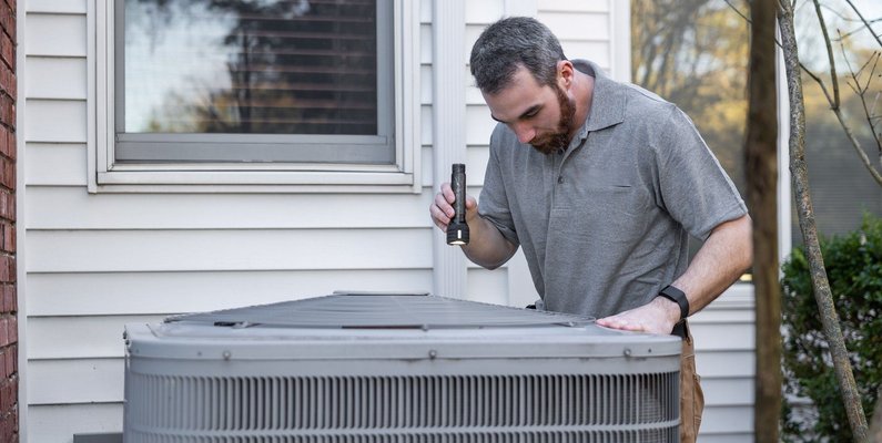 An HVAC technician inspecting an outdoor condenser unit with a flashlight