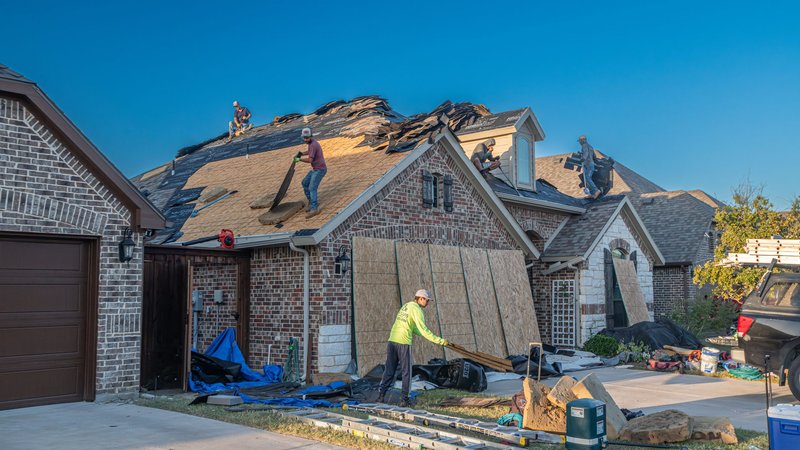 A roofing crew tearing off an old roof on a brick home, with the decking exposed and the walls sheeted for protection